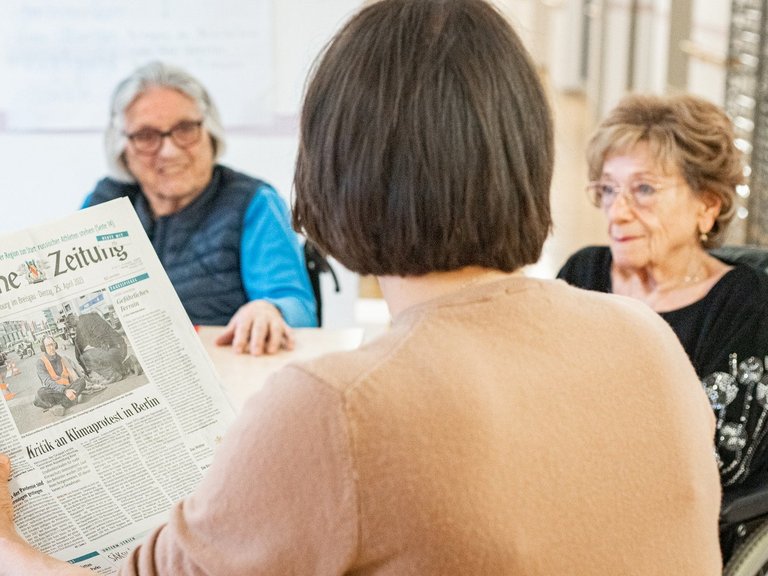 Eine Betreuerin liest Seniorinnen aus der Zeitung vor