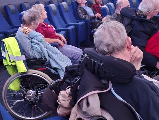 Eine Gruppe älterer Menschen sitzt in blauen Reihen, vermutlich in einem Bus oder einer Wartehalle. Mehrere Personen befinden sich in Rollstühlen, darunter eine Person mit einer auffälligen gelben Warnweste.